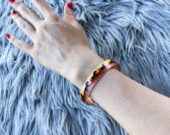 Op de afbeelding: Een gouden armband met een kleurrijk ontwerp met silhouetten van paarden. De armband zit om een pols.