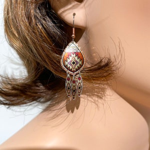 May include: A pair of dangle earrings with a teardrop-shaped pendant featuring a geometric pattern in shades of red, orange, white, and black. The pendant is accented with three smaller teardrop-shaped charms with a similar pattern.