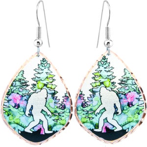 May include: A pair of silver dangle earrings with a teardrop-shaped design. Each earring features a watercolor-style image of a white silhouette of Bigfoot walking through a forest with green and pink trees.