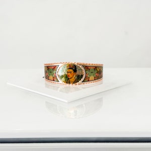 May include: Copper-toned cuff bracelet featuring a portrait of Frida Kahlo. The central image is framed by an oval border, surrounded by a floral and foliage design. The bracelet is displayed on a white surface.
