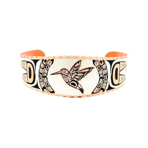 May include: A copper cuff bracelet featuring a central hummingbird motif and stylized geometric patterns in black and white. The metal band has a textured, scalloped edge and an open back design, showcasing a polished finish with intricate engraved details.