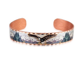 May include: A copper cuff bracelet featuring a central engraved design of a soaring eagle with outstretched wings. The background depicts a detailed mountain landscape with evergreen trees in shades of silver, grey, and dark green.