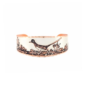May include: A wide copper cuff bracelet featuring a white enamel surface with a detailed black etched illustration of a roadrunner bird in a desert landscape with cacti and rock formations, finished with a textured copper edge along the top and bottom.