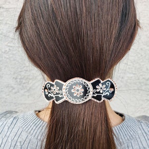 Puede incluir: Pinza para el pelo negra con un diseño floral blanco. La pinza tiene tres secciones, cada una con un patrón floral diferente. La pinza se lleva en el pelo de una mujer.