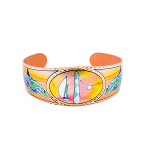 May include: Copper cuff bracelet with a coastal design. The bracelet features a central oval with a sailboat, lighthouse, and seagull. Dolphins are on either side. The background has orange, yellow, and pink hues.