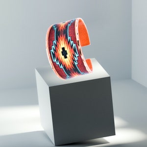 May include: A copper cuff bracelet with a colorful geometric pattern in shades of red, orange, blue, and black. The bracelet is on a white surface.