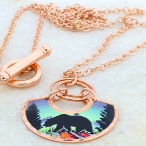 May include: A copper necklace with a pendant featuring a black bear silhouette against a colorful forest scene. The necklace has a toggle clasp.