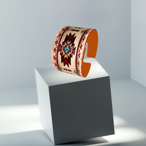 May include: A wide leather cuff bracelet with a colorful geometric pattern in shades of red, orange, blue, and white. The bracelet is on a white surface.