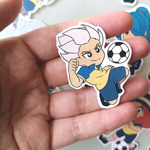 May include: A cartoon sticker of a soccer player with white hair, a yellow and blue jersey, and a soccer ball. The sticker is on a hand.