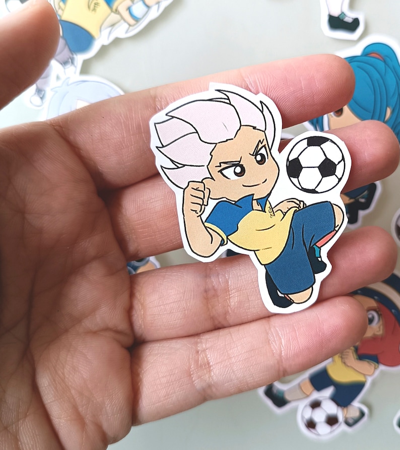May include: A cartoon sticker of a soccer player with white hair, a yellow and blue jersey, and a soccer ball. The sticker is on a hand.