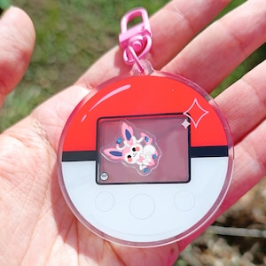 May include: A pink and red Pokemon character keychain with a clear plastic charm featuring a pink Eevee character. The keychain has a silver metal ring and a pink plastic clip.