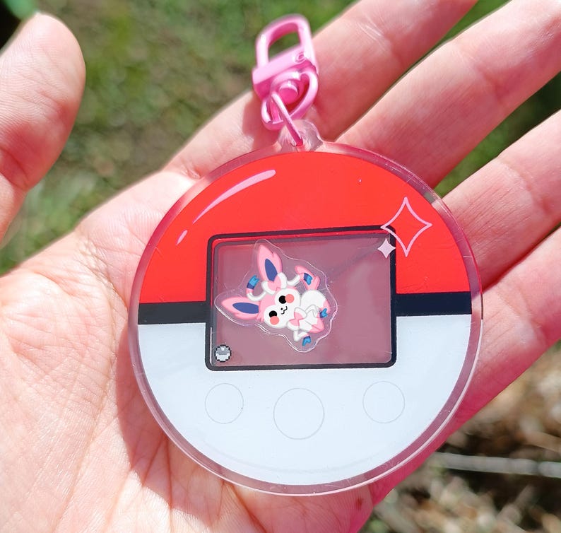 May include: A pink and red Pokemon character keychain with a clear plastic charm featuring a pink Eevee character. The keychain has a silver metal ring and a pink plastic clip.