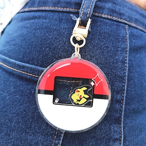 May include: A round, clear plastic keychain with a red, white, and black Pokeball design. A yellow Pikachu character is visible inside the keychain.