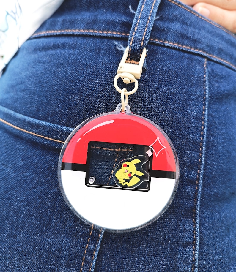 May include: A round, clear plastic keychain with a red, white, and black Pokeball design. A yellow Pikachu character is visible inside the keychain.