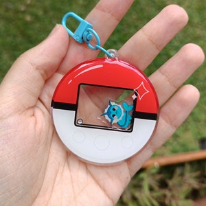 May include: A red, white, and blue Pokemon keychain with a clear plastic window featuring a blue and white Pokemon character. The keychain has a blue metal ring.