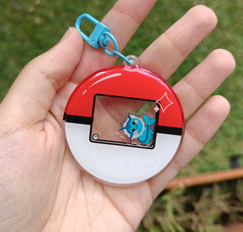 May include: A red, white, and blue Pokemon keychain with a clear plastic window featuring a blue and white Pokemon character. The keychain has a blue metal ring.