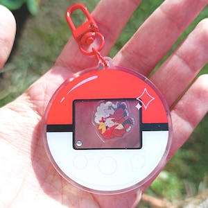 May include: A red and white Pokemon character keychain with a clear plastic casing. The keychain features a red and white Pokeball design with a clear window showing a cartoon image of a red and white Pokemon character.