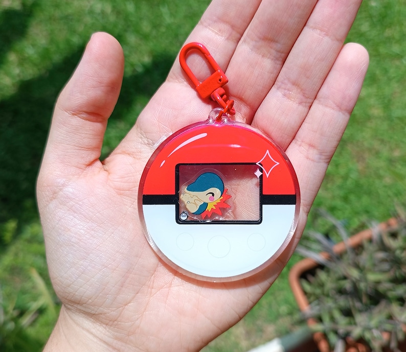 May include: A red, white, and black Pokemon keychain with a clear plastic cover. The keychain features a small, cartoon Pokemon character inside the cover.