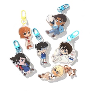 Op de afbeelding: Set van zeven kleurrijke anime-personage sleutelhangers met blauwe en gele metalen ringen. De personages zijn getekend in chibi-stijl en omvatten een detective, een meisje met bruin haar en een jongen met bruin haar.