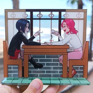 May include: A decorative anime-style diorama featuring two figures seated at a table. One figure has black hair and wears a black jacket and red skirt. The other figure has pink hair and wears a white shirt and pink skirt. The scene is set in a cafe with a window overlooking the ocean.