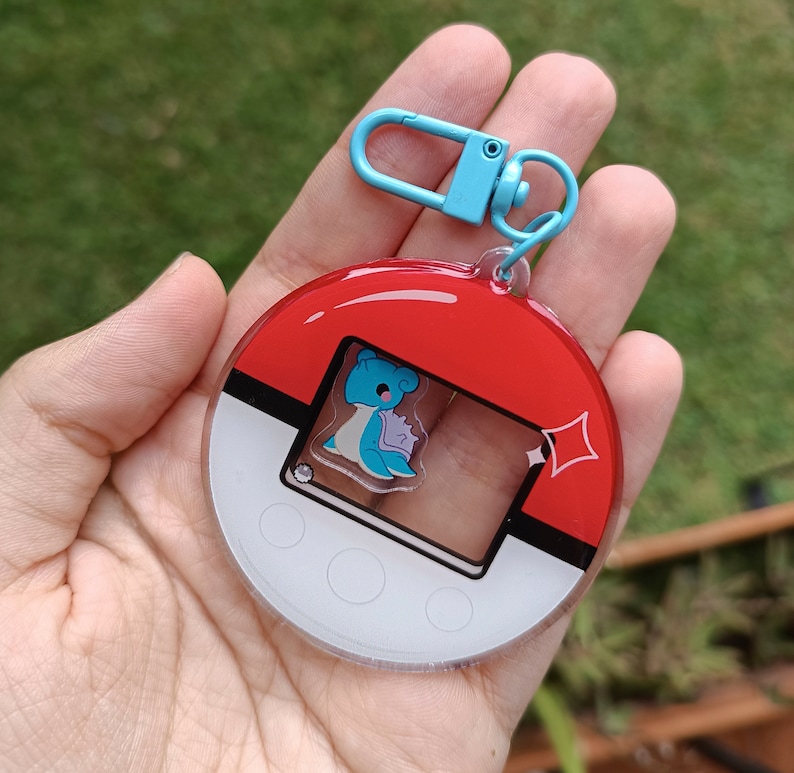 May include: A red, white, and blue Pokemon-themed keychain with a blue character inside a clear window. The keychain has a silver ring and a blue clip.
