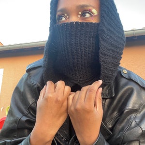 May include: A person wearing a black leather jacket with a black knit hooded scarf pulled up over their face. The person has colorful makeup around their eyes.