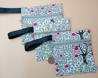 May include: Three square zippered pouches featuring a white fabric with a repeating pattern of black tree branches, grey leaves, red apples, and green pears. Each pouch includes a grey zipper closure and a black fabric wrist strap attached to the side.