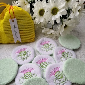 Op de afbeelding: Een set van 10 herbruikbare groene en witte make-up remover pads met geborduurde distels. De pads zitten in een gele mesh-tas met een wit label waarop "Handmade" staat.