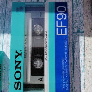 Puede incluir: Una caja de cinta de casete azul y turquesa con el texto "SONY" en blanco, "EXCELLENT FIDELITY" en blanco, "Japan" en blanco, "HIGH RESISTANCE TO HEAT" en blanco, "TYPE I (NORMAL) POSITION" en blanco, "COMPACT CASSETTE/CASSETTE COMPACTE" en blanco y "EF90" en blanco. La caja tiene una ventana de plástico transparente que muestra la cinta de casete en el interior.