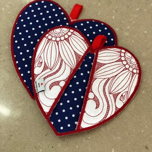 Op de afbeelding: Twee hartvormige ovenwanten. Eén is donkerblauw met witte stippen en de andere is wit met een rood bloemenmotief. Beide hebben rode stiksels rond de randen.