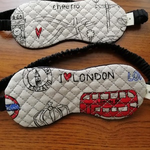 以下が含まれることがあります： 黒いゴムバンドが付いた、2つのグレーのキルティングスリープマスク。1つのマスクには赤いハートと「cheerio」の文字、もう1つは赤い2階建てバスのイラストとともに「I ❤️ LONDON」と書かれています。どちらもロンドンをテーマにしたスケッチが描かれています。