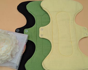 Könnte beinhalten: Drei wiederverwendbare Stoffbinden in Schwarz, Olivgrün und Hellgelb, jeweils mit konturierter Form, seitlichen Flügeln und Druckknöpfen. Ein weißer Wäschenetzbeutel mit einer befestigten weißen Stoffblume befindet sich links neben den Binden.
