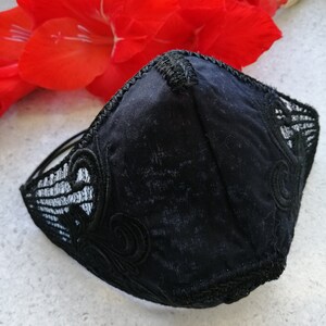 May include: A black face mask featuring a central seam and intricate black lace overlays on the sides. The mask has a structured, contoured shape with decorative embroidery details along the edges and thin black elastic ear loops for secure attachment.