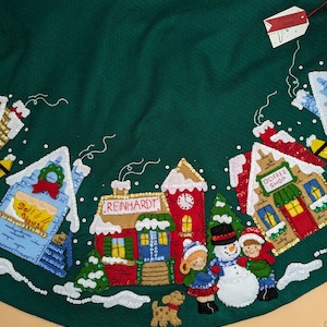 Pode incluir: Uma saia de árvore de Natal de feltro verde com uma cena de vila de inverno. A saia apresenta uma escola, uma cafeteria, uma casa de bonecas, um boneco de neve e duas crianças. A saia é decorada com lantejoulas e contas.