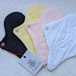 Pode incluir: Uma coleção de pensos higiénicos de tecido reutilizáveis em preto, amarelo, rosa e branco. Cada penso tem uma forma única e é fixado com um fecho de pressão. Uma etiqueta com a marca "Felinia Di" está anexada.