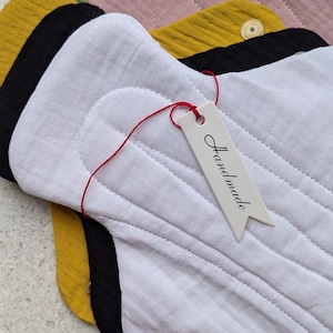 May include: A stack of reusable cloth menstrual pads in white, black, mustard yellow, dusty pink, and olive green. The top white pad features a small, clear plastic snap button and a white paper tag tied with red string that says Handmade.