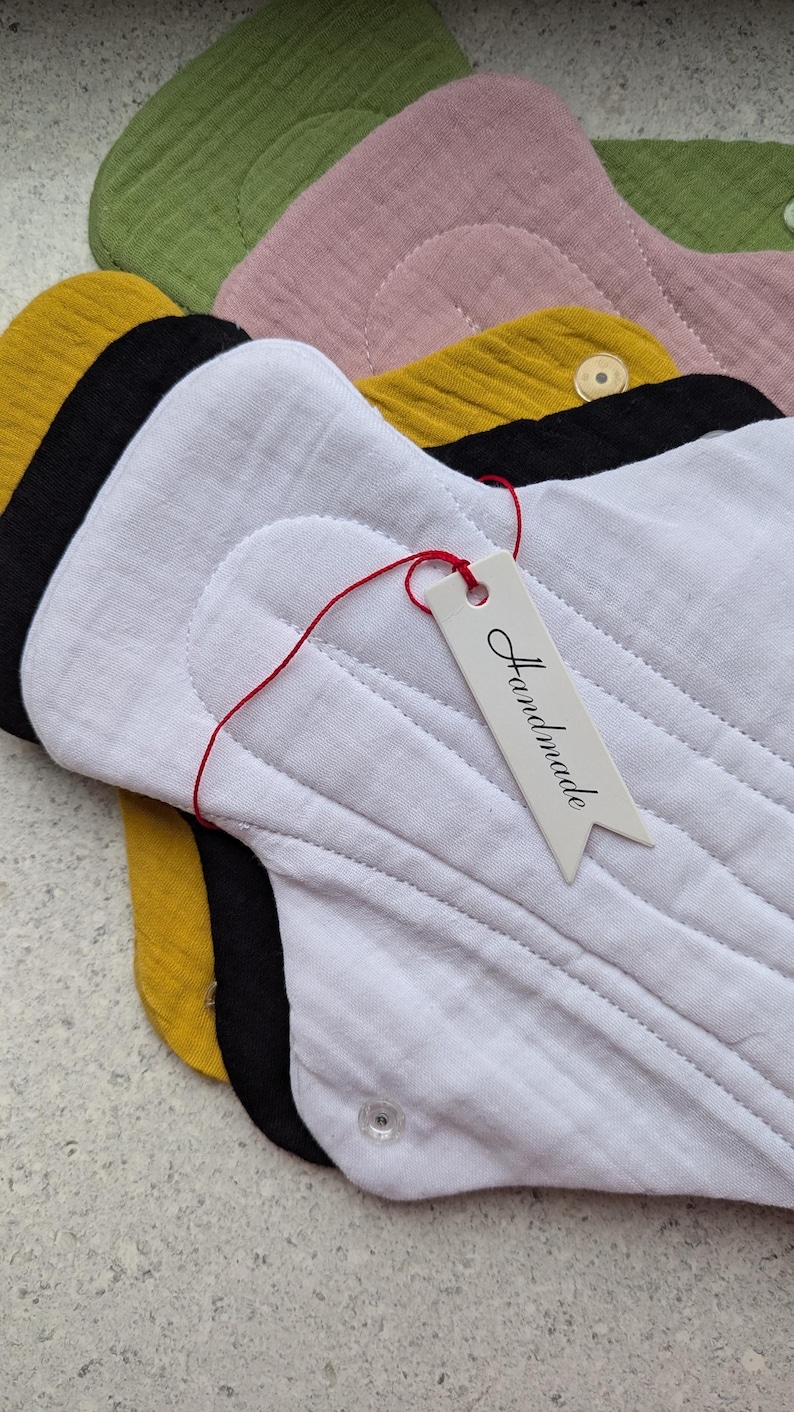 May include: A stack of reusable cloth menstrual pads in white, black, mustard yellow, dusty pink, and olive green. The top white pad features a small, clear plastic snap button and a white paper tag tied with red string that says Handmade.