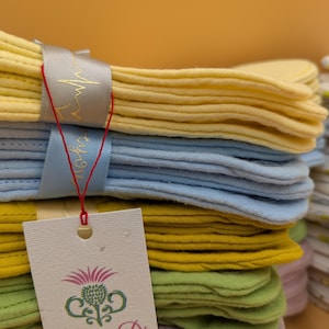 May include: A stack of folded fabric cloths in pastel yellow, light blue, mustard yellow, sage green, and soft pink. A grey ribbon with a gold heartbeat graphic wraps around the stack. A white rectangular tag features a thistle logo and the text FotiniaDi.