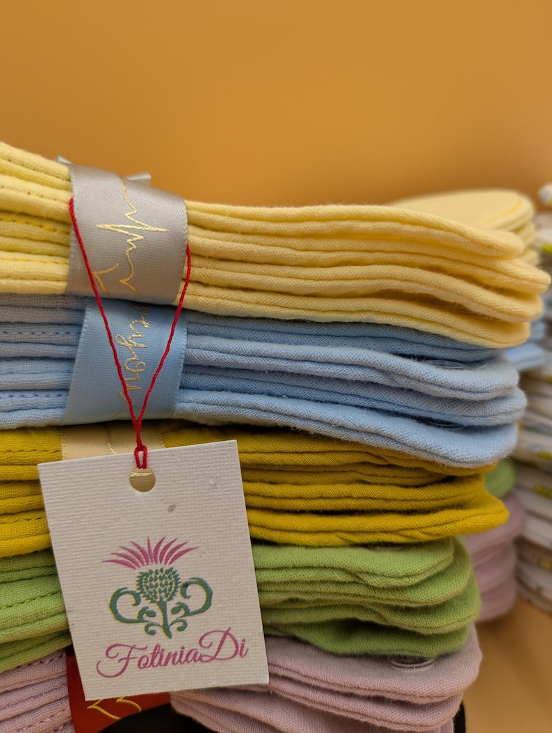 May include: A stack of folded fabric cloths in pastel yellow, light blue, mustard yellow, sage green, and soft pink. A grey ribbon with a gold heartbeat graphic wraps around the stack. A white rectangular tag features a thistle logo and the text FotiniaDi.