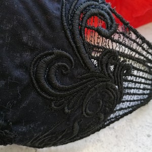 May include: A black fabric garment featuring intricate black embroidery with swirling, ornate patterns. The design includes a section of delicate, open-work lace detailing along the edge, contrasting with the solid black textile base.