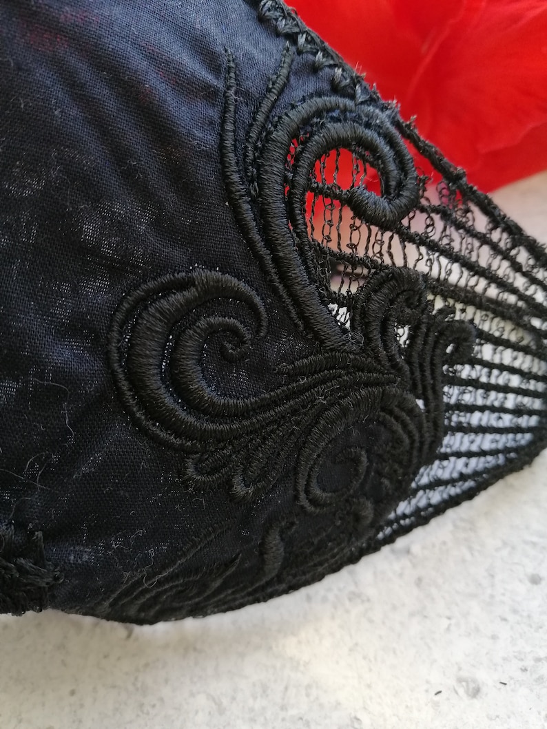 May include: A black fabric garment featuring intricate black embroidery with swirling, ornate patterns. The design includes a section of delicate, open-work lace detailing along the edge, contrasting with the solid black textile base.