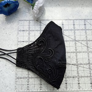 May include: A black face mask featuring intricate black embroidery and lace side panels, positioned on a clear ruler showing a width of approximately 15 cm and a height of approximately 10 cm, with black elastic ear loops attached.