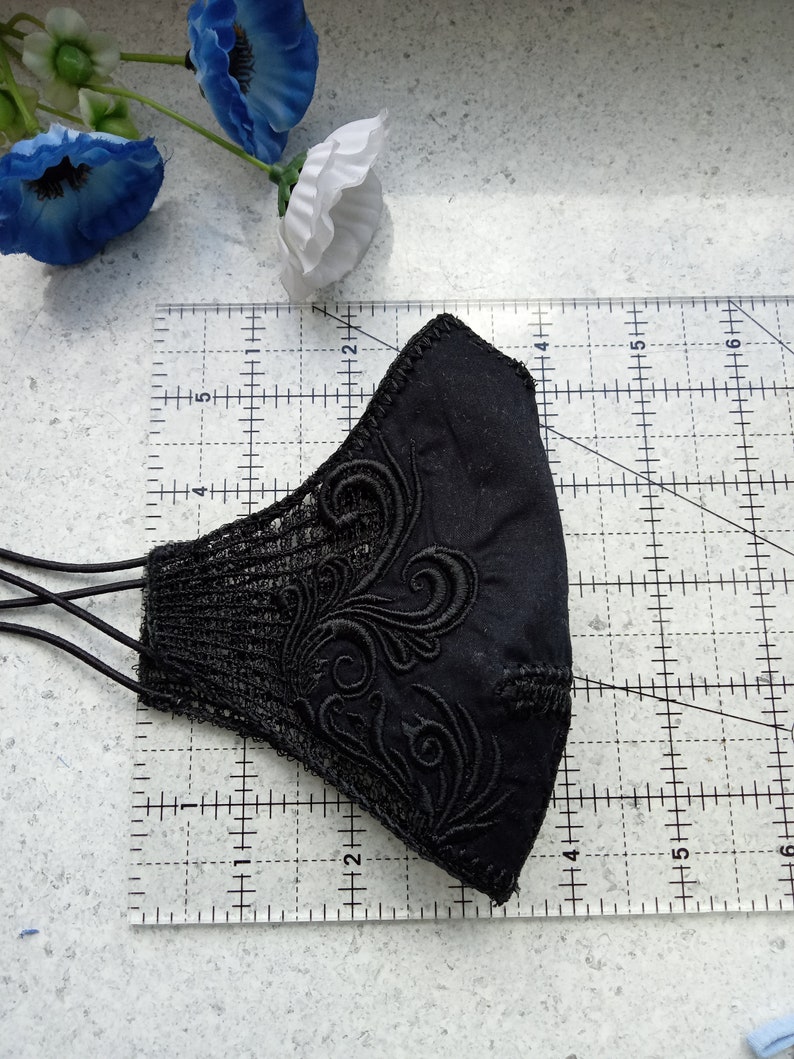 May include: A black face mask featuring intricate black embroidery and lace side panels, positioned on a clear ruler showing a width of approximately 15 cm and a height of approximately 10 cm, with black elastic ear loops attached.