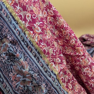 Può includere: Primo piano di un tessuto bordeaux con un motivo floreale nei toni dell'arancione, dell'oro e del crema. Un bordo blu scuro con un motivo floreale e finiture dorate corre lungo il bordo. Il tessuto è drappeggiato, mostrando la sua consistenza e il suo design.