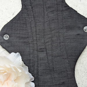 May include: A black reusable cloth menstrual pad featuring a textured fabric surface with vertical stitched channels for structure. The pad includes two clear plastic snap fasteners on the side wings to secure it in place during use.