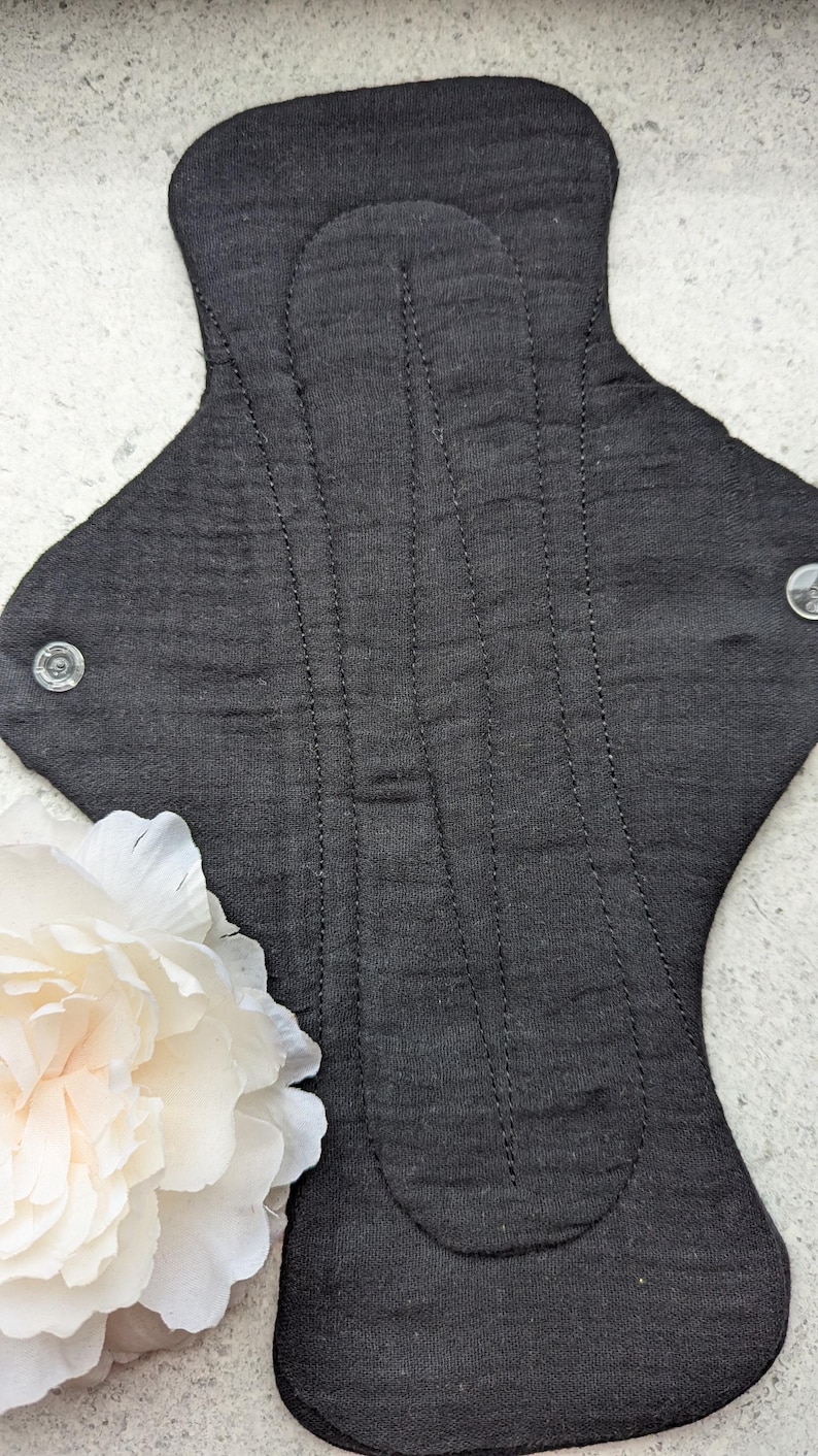 May include: A black reusable cloth menstrual pad featuring a textured fabric surface with vertical stitched channels for structure. The pad includes two clear plastic snap fasteners on the side wings to secure it in place during use.