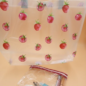 May include: A clear tote bag featuring a repeating red tomato pattern with grey handles and a silver snap closure, accompanied by a smaller clear zippered pouch containing various cosmetic items including tubes and a small blue bottle.