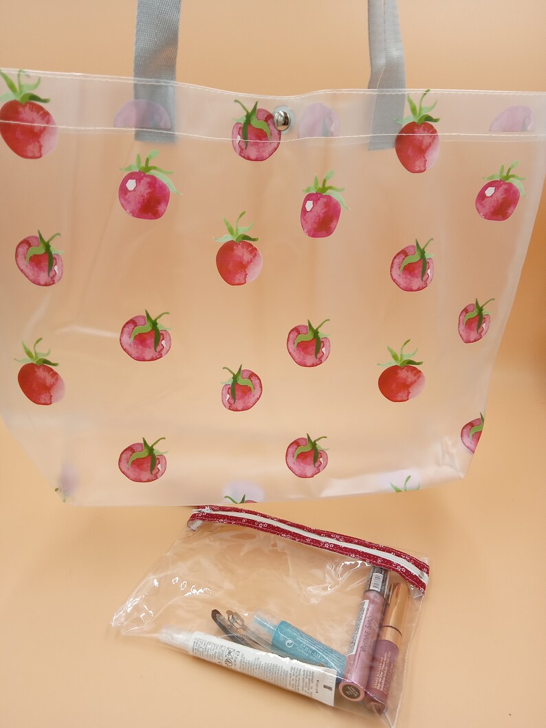 May include: A clear tote bag featuring a repeating red tomato pattern with grey handles and a silver snap closure, accompanied by a smaller clear zippered pouch containing various cosmetic items including tubes and a small blue bottle.