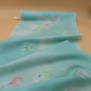 May include: A rolled-up, light teal fabric with a floral pattern. The fabric has a watercolor effect with white, pink, and yellow flowers. The background has a subtle wave-like texture and small white dots.
