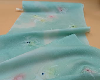 May include: A folded length of light mint green fabric featuring a subtle, flowing wave pattern and scattered, soft-focus floral motifs in shades of pale pink, white, and yellow, accented with tiny white dots across the surface.